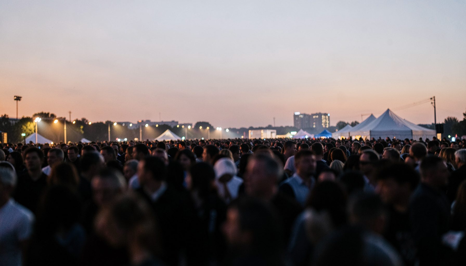 A wide atmospheric photograph of a large public gathering at dusk, with soft amber and navy tones and blurred crowd silhouettes